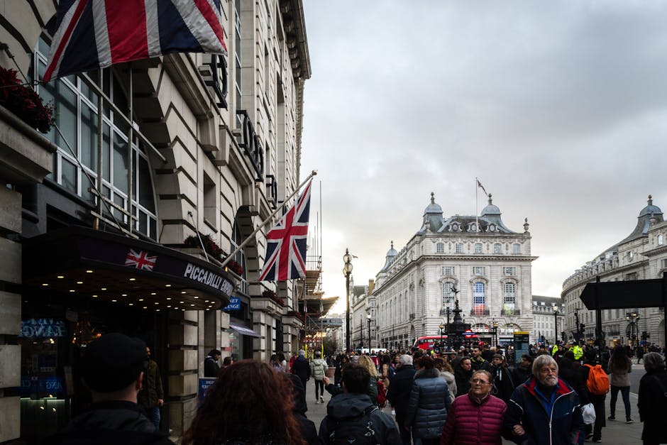 A busy urban scene outside Piccadilly Circus in London, featuring a crowd of pedestrians walking along the sidewalk in front of a building with Union Jack flags and a sign reading 'Piccadilly.' The st