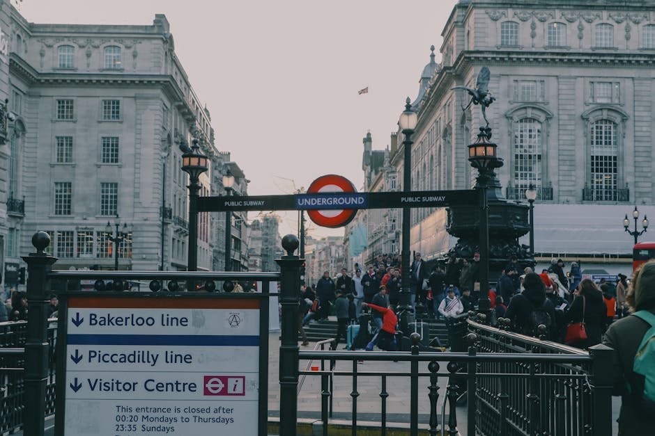 A busy urban scene outside Piccadilly Circus in London, featuring a crowd of pedestrians walking along the sidewalk in front of a building with Union Jack flags and a sign reading 'Piccadilly.' The st