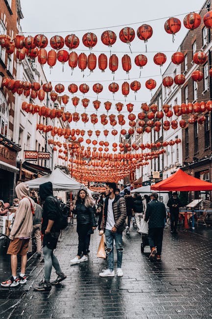 A bustling outdoor street scene in Carnaby Street with numerous red paper lanterns hanging overhead in evenly spaced rows, creating a festive atmosphere. The street is paved with dark, wet cobblestone